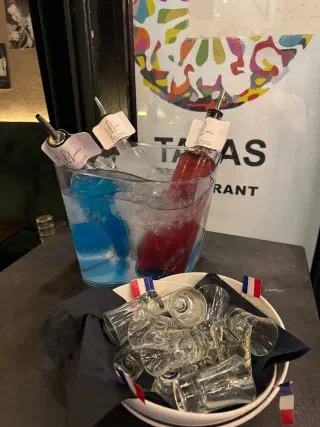Ice bucket with bottles of blue and red cocktails; beside it, a bowl of shot glasses with small French flags