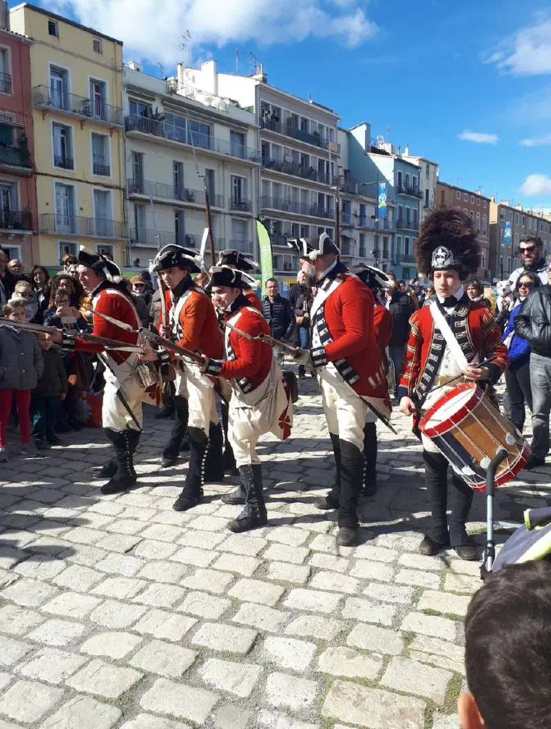 Reenactors in red uniforms with muskets and a drummer on the cobbled quay, surrounded by a festival crowd