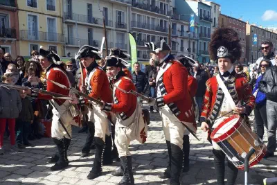 Reenactors in red uniforms with muskets and a drummer on the cobbled quay, surrounded by a festival crowd