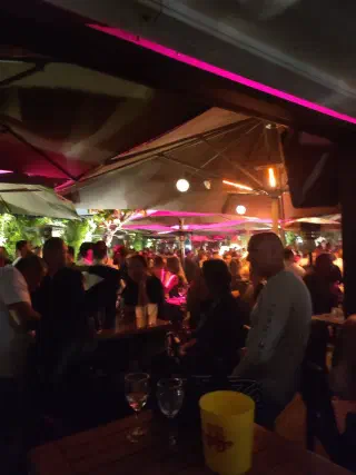 Crowded evening terrace under parasols lit in pink, with wine glasses on a table in the foreground