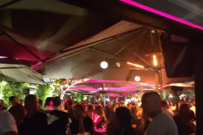 Crowded evening terrace under parasols lit in pink, with wine glasses on a table in the foreground