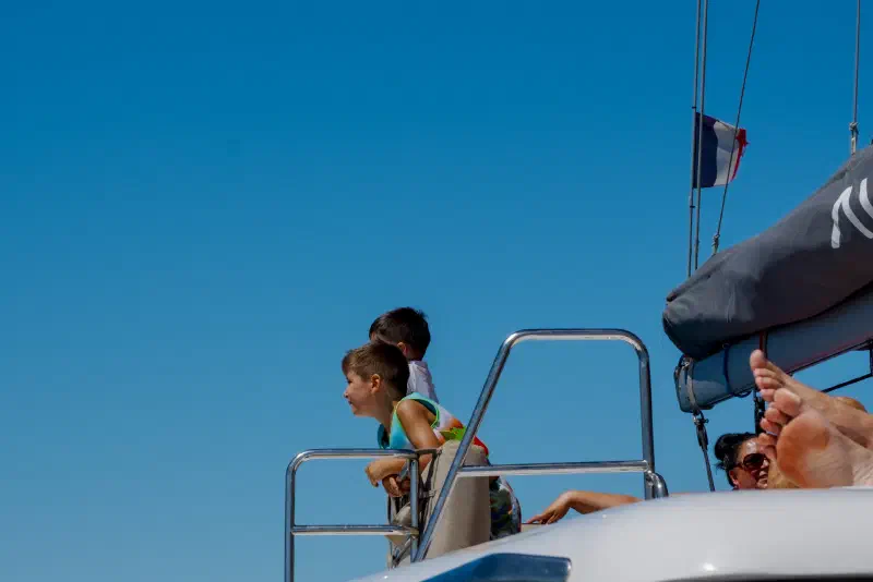 Two boys at the deck rail of a catamaran look out to sea, a French flag above them