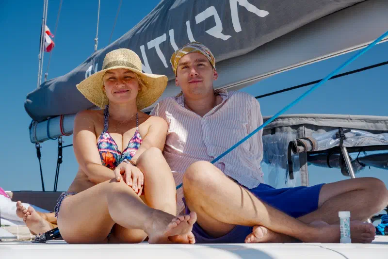 A couple sits on the catamaran's foredeck under the covered mainsail against a blue sky