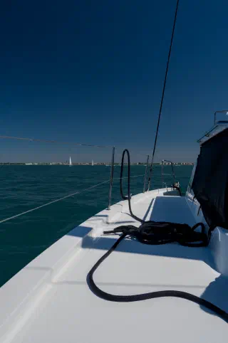 White side deck of the catamaran with a mooring line; a low shoreline with houses and sails beyond