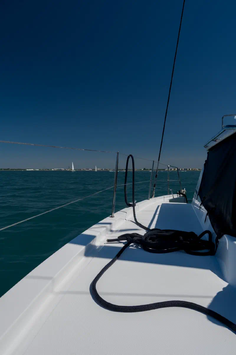 White side deck of the catamaran with a mooring line; a low shoreline with houses and sails beyond