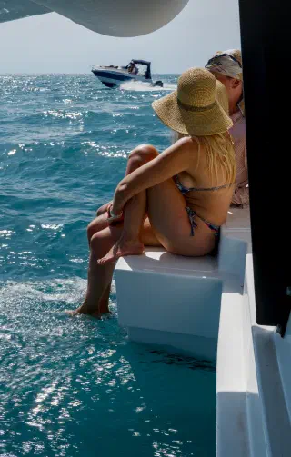 Two people sit on the catamaran's stern step, feet in the water, as a motorboat passes by