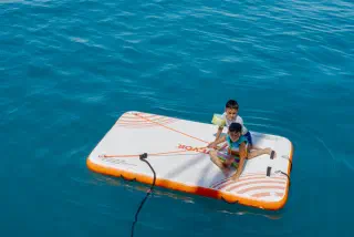 Two children on an inflatable platform in the middle of the turquoise sea, seen from the boat