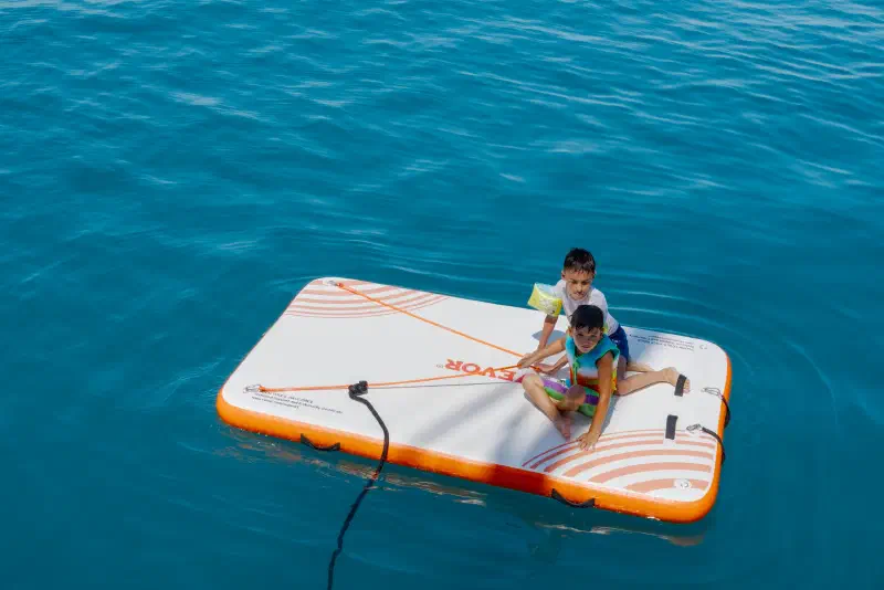 Two children on an inflatable platform in the middle of the turquoise sea, seen from the boat