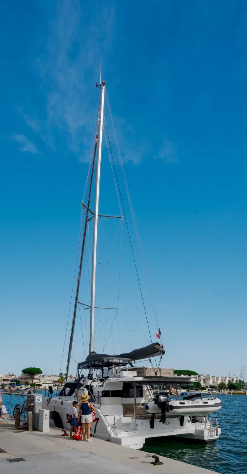 Catamaran with a tall mast at the quay in the morning; an adult and a child with bags beside it