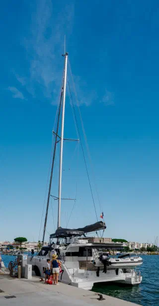 Catamaran with a tall mast at the quay in the morning; an adult and a child with bags beside it