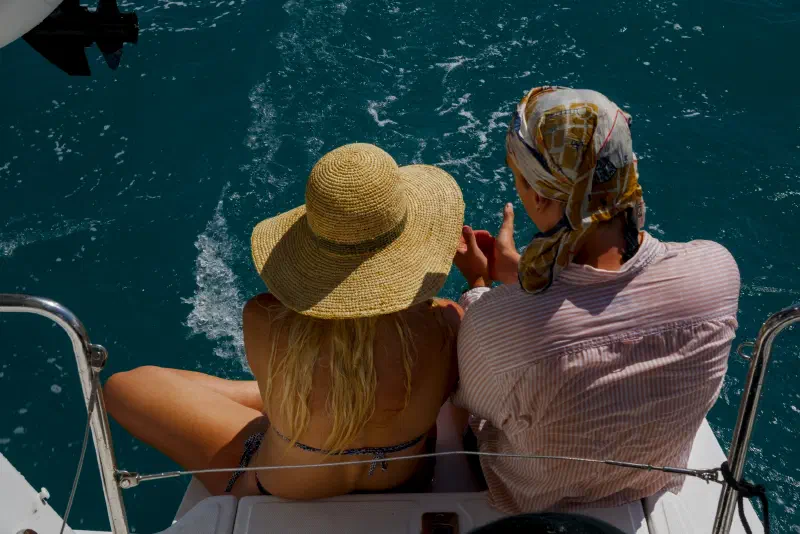 Two people sit at the stern of a catamaran at sea, watching the foamy wake below