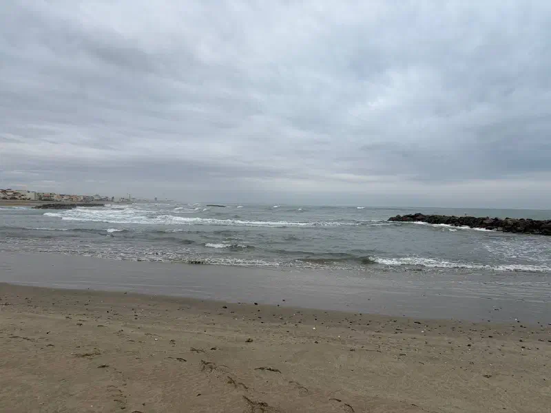 An empty sandy beach on an overcast day, with a choppy grey sea and a rocky breakwater near the shore.