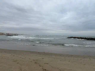 An empty sandy beach on an overcast day, with a choppy grey sea and a rocky breakwater near the shore.