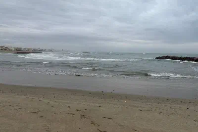 An empty sandy beach on an overcast day, with a choppy grey sea and a rocky breakwater near the shore.
