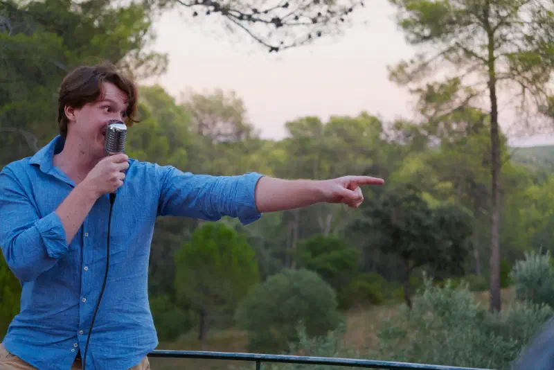 A singer with a vintage microphone on the terrace points towards the guests, pines and a pink sunset sky behind