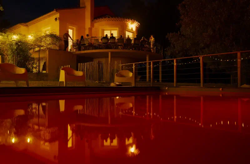 The villa at night: guests at a long table on the round terrace, a red-lit pool and string lights below