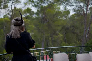 A guest in a black dress and feathered headband stands with her back to the camera at the terrace railing, looking at the pine park