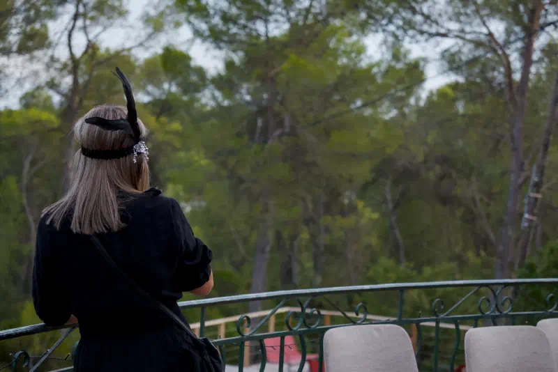 A guest in a black dress and feathered headband stands with her back to the camera at the terrace railing, looking at the pine park