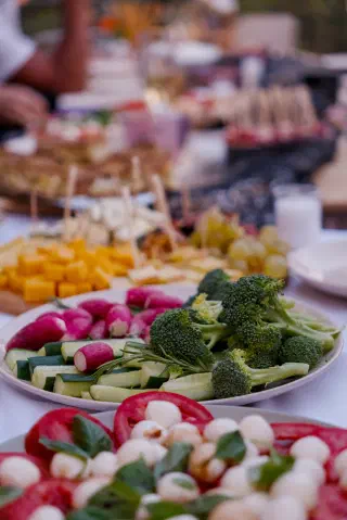 A plate of broccoli, cucumber and radishes, with a cheese board and grapes behind it on the long table