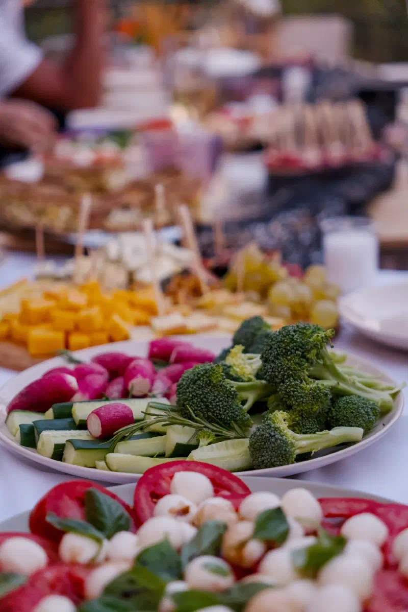 A plate of broccoli, cucumber and radishes, with a cheese board and grapes behind it on the long table