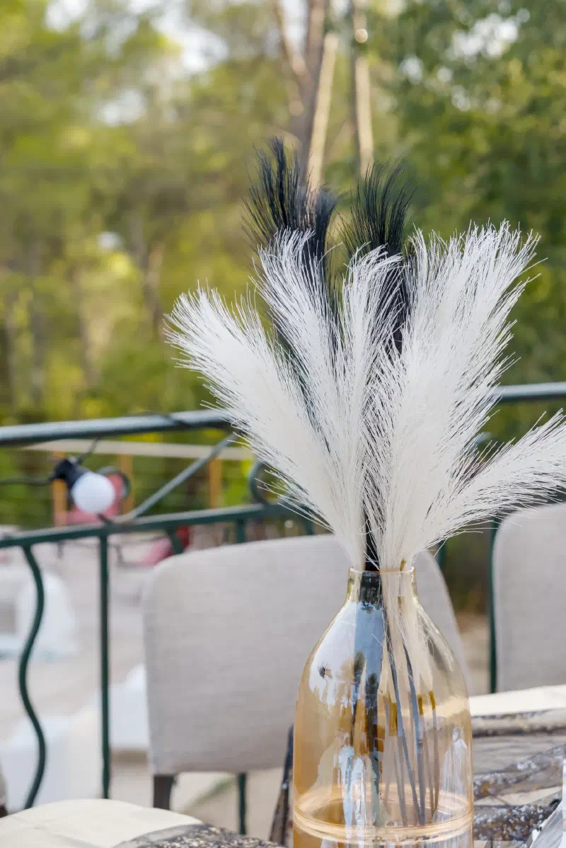 White and black pampas grass in a glass vase on the terrace table, pines and string lights beyond the railing