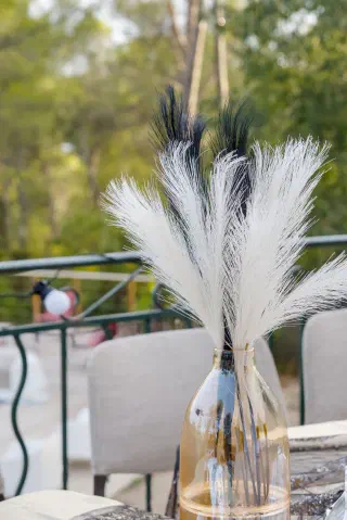 White and black pampas grass in a glass vase on the terrace table, pines and string lights beyond the railing