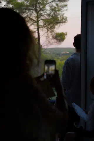 Silhouette of a guest photographing the sunset over the park from the villa balcony, a guest in a white shirt beside her