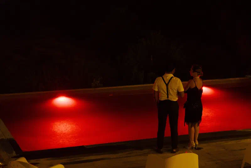 A couple in 1920s style, braces and a fringed dress, stand with their backs to the camera by the red-lit pool at night