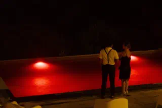 A couple in 1920s style, braces and a fringed dress, stand with their backs to the camera by the red-lit pool at night