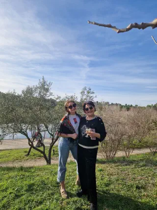 Two women posing by an olive tree at the water's edge, one holding a glass