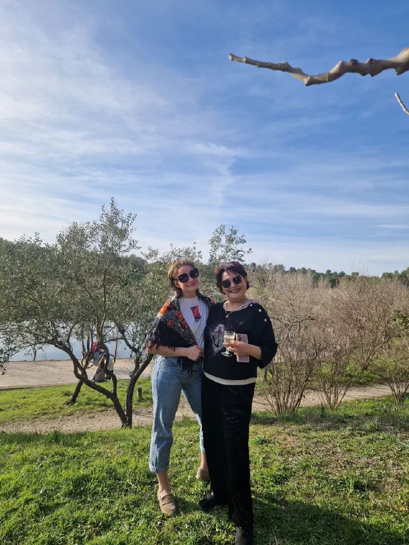 Two women posing by an olive tree at the water's edge, one holding a glass