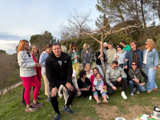 Evening group photo around a young tree on a lawn; a girl with a plush unicorn sits in front