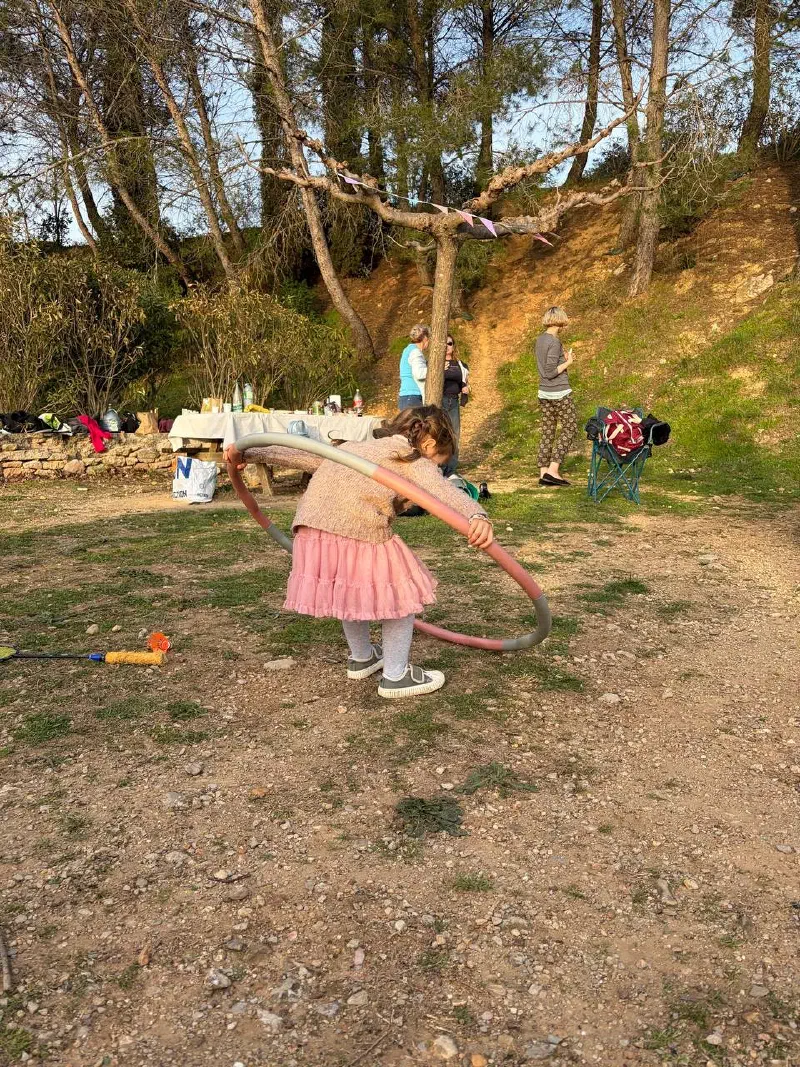 A little girl in a pink dress spins a hula hoop in evening light; a laid table and guests under the pines behind her