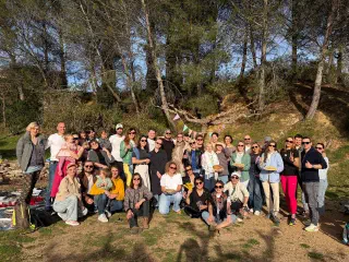 A big group posing with plates of pancakes under pine trees in a sunny park, with bunting strung on a tree