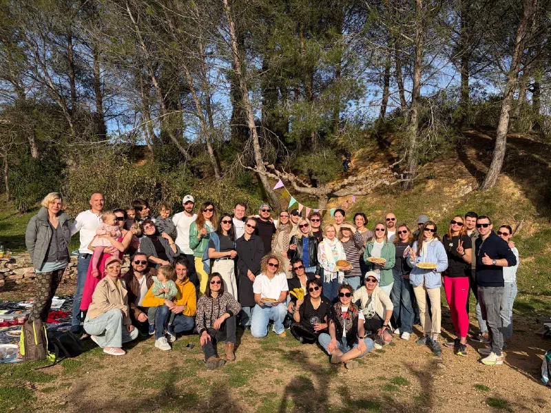 A big group posing with plates of pancakes under pine trees in a sunny park, with bunting strung on a tree