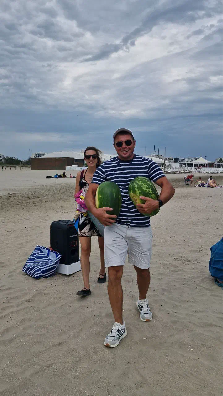 A man in a striped T-shirt carries two watermelons under his arms across a sandy beach, past a speaker and bags.