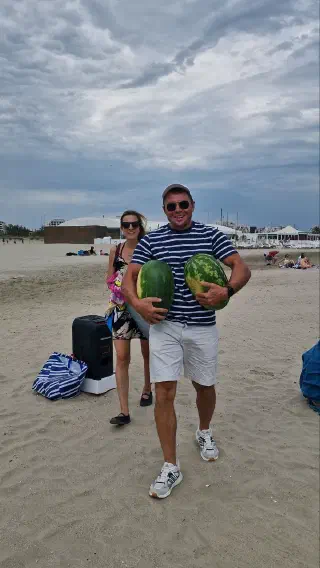 A man in a striped T-shirt carries two watermelons under his arms across a sandy beach, past a speaker and bags.