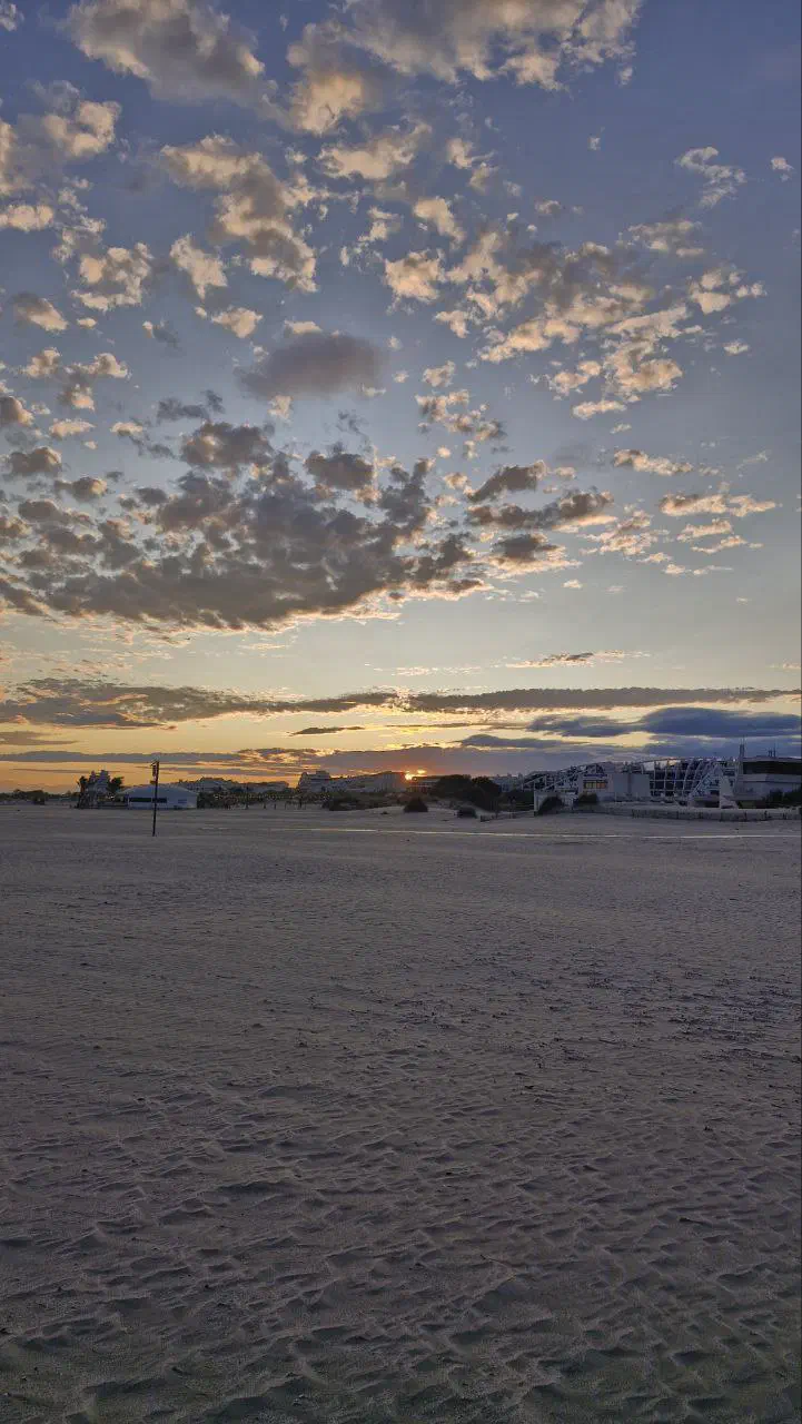 Sunset over an empty sandy beach, the sun dipping behind buildings on the horizon under scattered clouds.