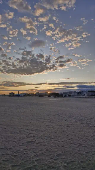 Sunset over an empty sandy beach, the sun dipping behind buildings on the horizon under scattered clouds.