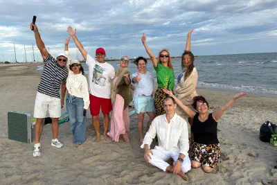 A cheerful group on a sandy beach with raised arms; the sea and yacht masts of a marina behind them.