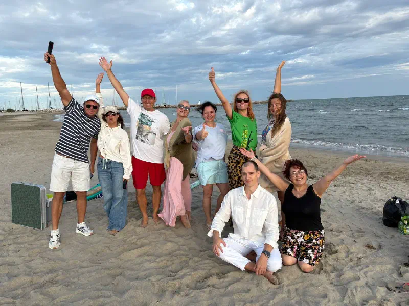 A cheerful group on a sandy beach with raised arms; the sea and yacht masts of a marina behind them.