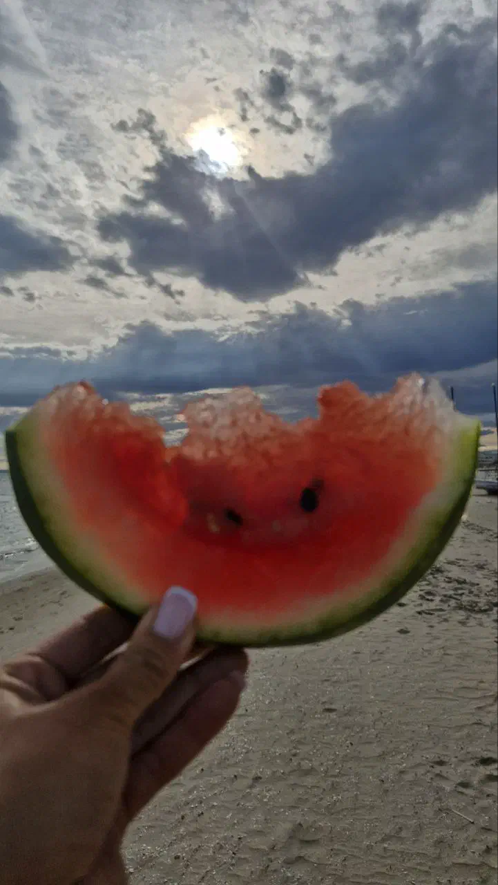 A hand holds a bitten slice of watermelon against a moody sky over the beach, sunlight breaking through clouds.