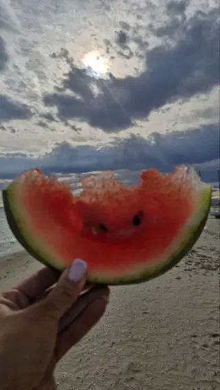 A hand holds a bitten slice of watermelon against a moody sky over the beach, sunlight breaking through clouds.