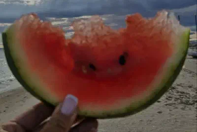 A hand holds a bitten slice of watermelon against a moody sky over the beach, sunlight breaking through clouds.