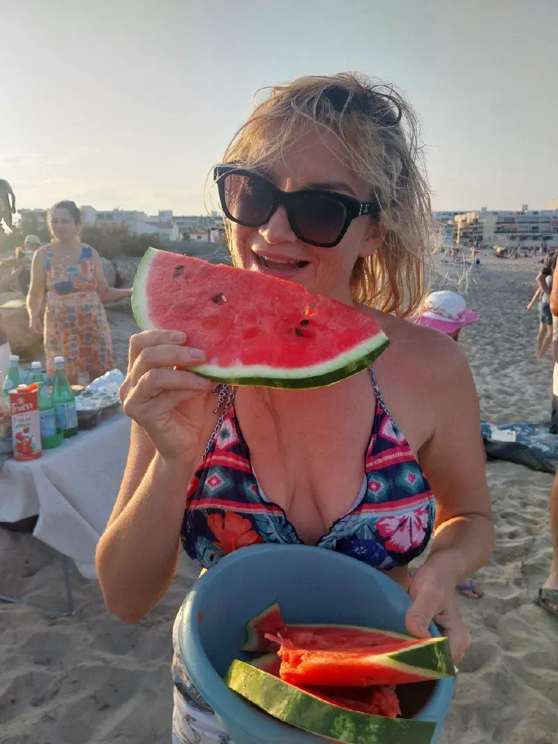 A participant holding a watermelon slice and a bowl of cut fruit by the shared table on an evening beach