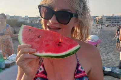 A participant holding a watermelon slice and a bowl of cut fruit by the shared table on an evening beach