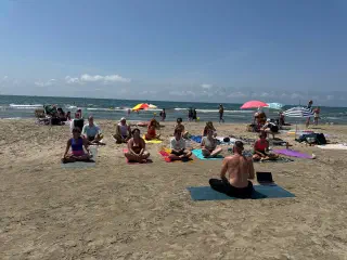 A group meditating cross-legged on mats on the sand, facing the instructor, with the sea and parasols behind