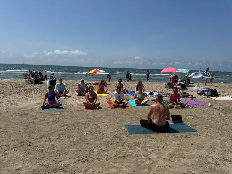 A group meditating cross-legged on mats on the sand, facing the instructor, with the sea and parasols behind