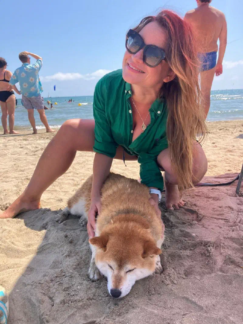 A smiling woman in a green shirt petting a ginger dog dozing on the sand by the sea