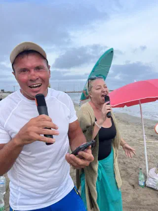 A man and a woman singing into microphones on the beach under an evening sky, a paddleboard and a pink parasol behind
